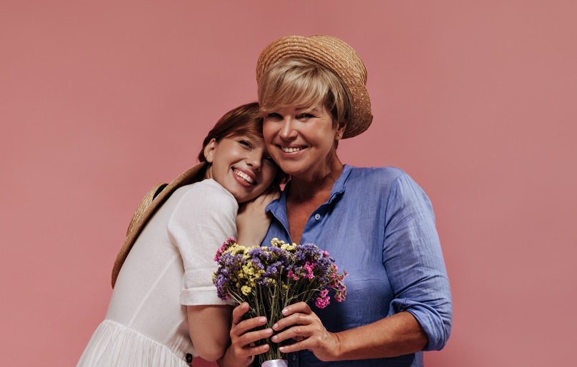 Trendy lady with blonde hairstyle in blue dress and straw hat smiling, holding colorful bouquet and posing with girl in white outfit on pink backdrop..