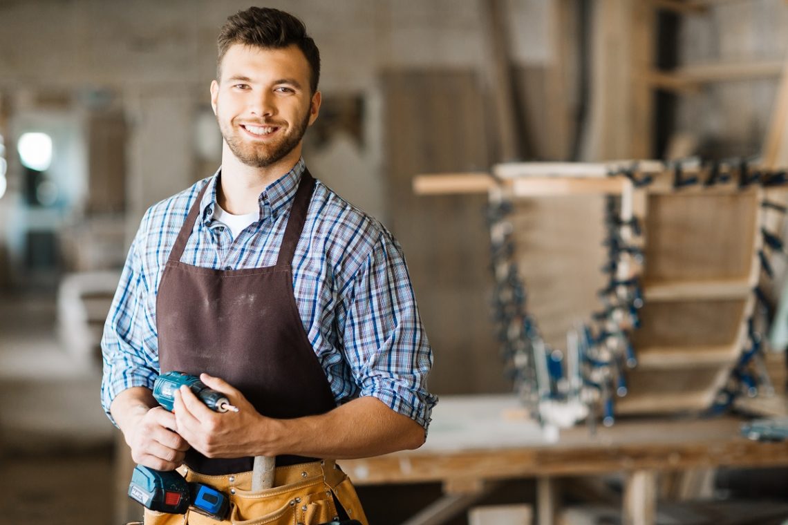 Waist-up portrait of smiling bearded craftsman with electric drill in hands standing in spacious workshop and looking at camera