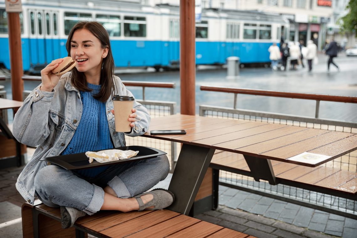 Attractive girl in casual style eats a burger with coffee sitting on the summer terrace of a cafe.