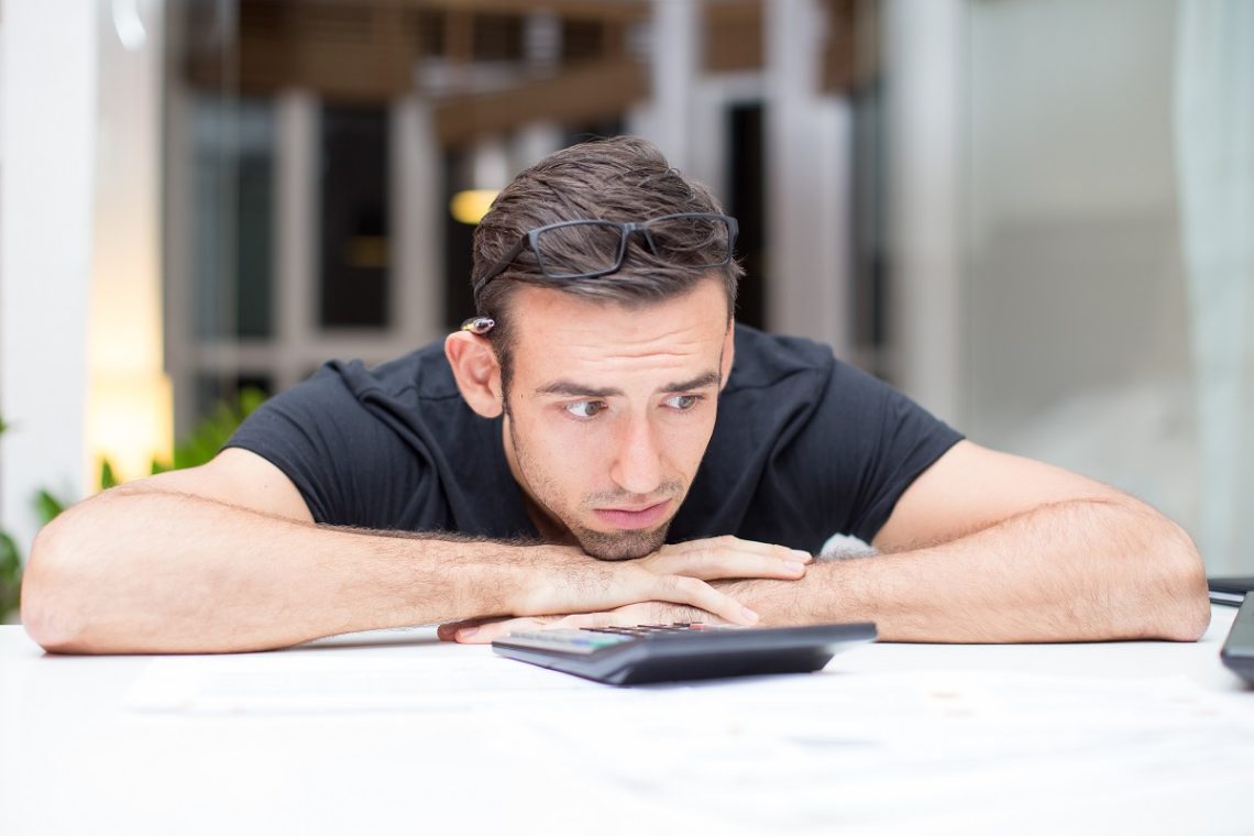 Closeup portrait of frustrated young handsome man lying on table with calculator in office