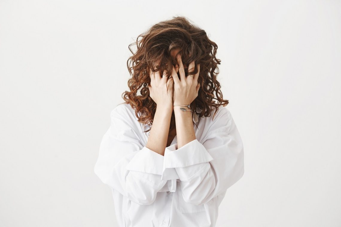 Studio portrait of tired or bothered woman hiding face in her curly hair and holding hands on it, expressing exhaustion or just being childish, standing over gray background. I am not seeing you.